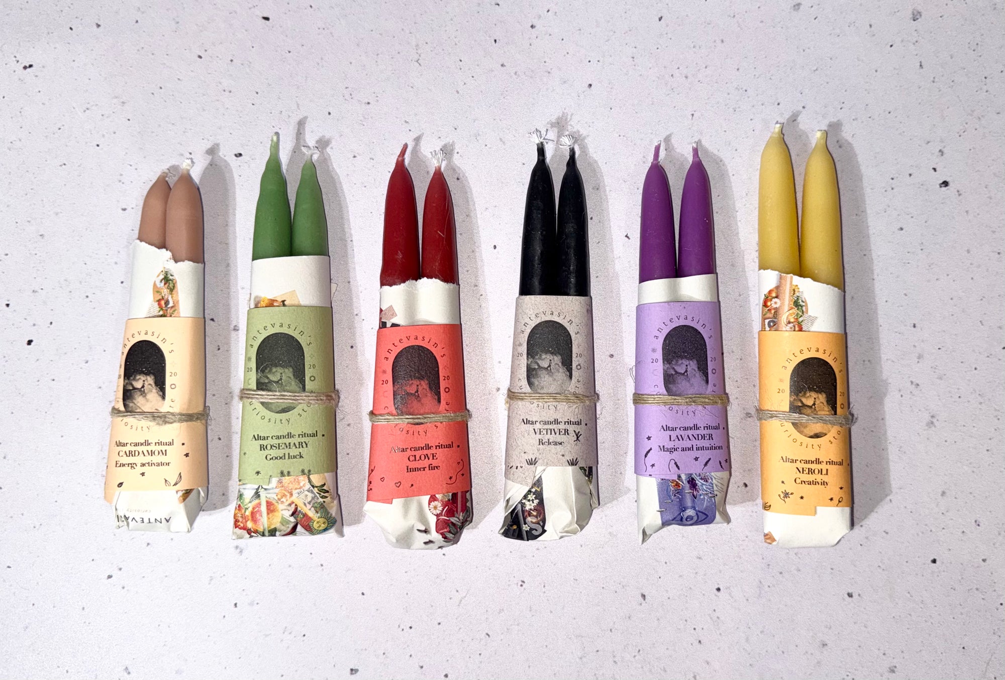 Six double taper candles in assorted colors are lined up horizontally on a light speckled background. The candles come in brown, green, red, black, purple, and yellow wax, each wrapped in a paper sleeve with a rustic twine bow and a vintage-style label.